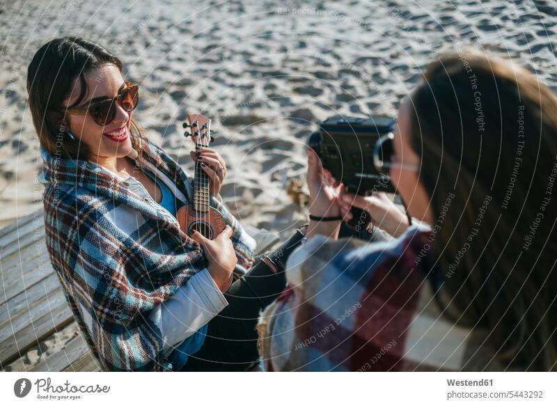 Smiling young woman playing ukulele while her friend recording with video camera female friends beach beaches mate friendship making music playing music
