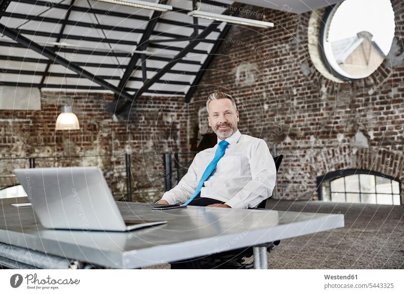 Portrait of confident businessman with laptop sitting at table in a loft Businessman Business man Businessmen Business men Seated Laptop Computers laptops