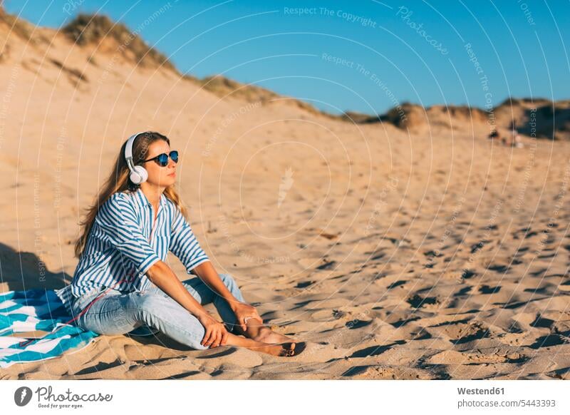 Portugal, Aveiro, woman sitting near beach dune listening music with headphones beach dunes Seated females women sand sandy hearing headset sand dune sand dunes