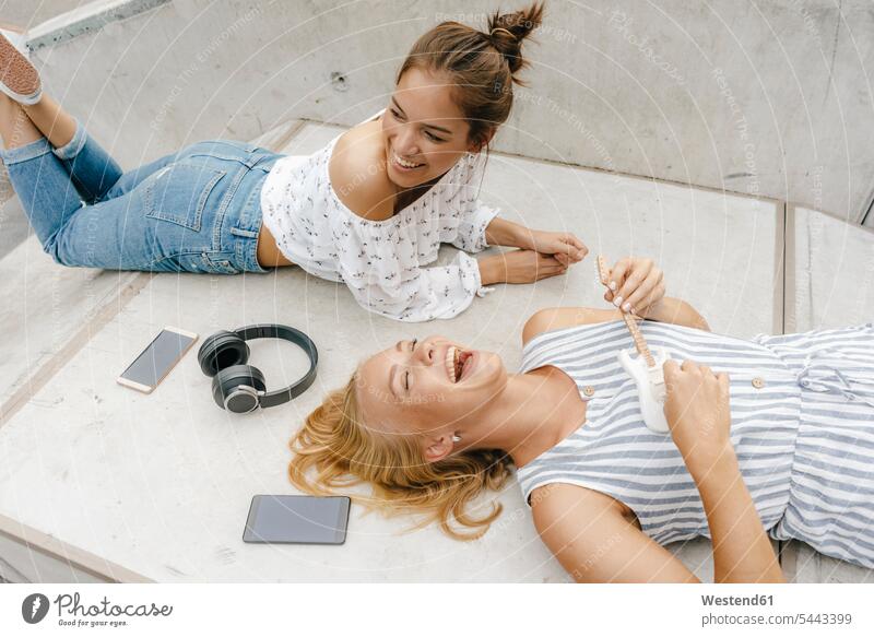 Two happy young women lying on ramp in a skatepark making music female friends woman females laying down lie lying down Skateboard Park skate park Ramp