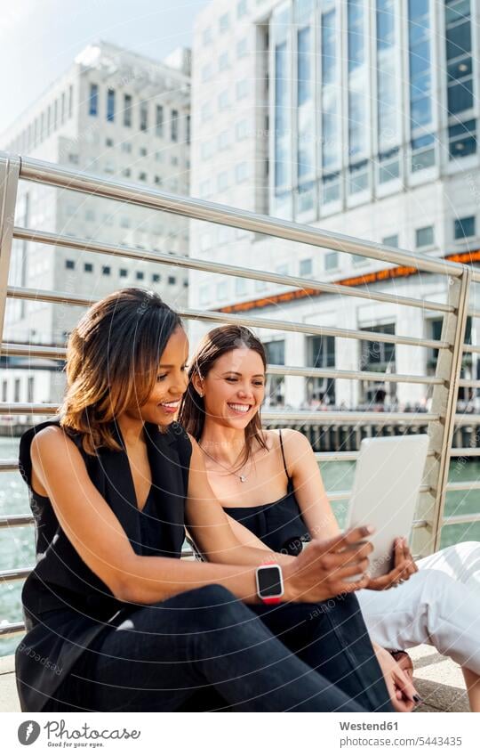 Two happy women sitting on a bridge sharing a tablet in the city Female Colleague smiling smile woman females digitizer Tablet Computer Tablet PC