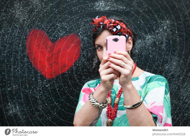Woman taking a selfie next to heart painted on the wall mobile phone mobiles mobile phones Cellphone cell phone cell phones woman females women telephones