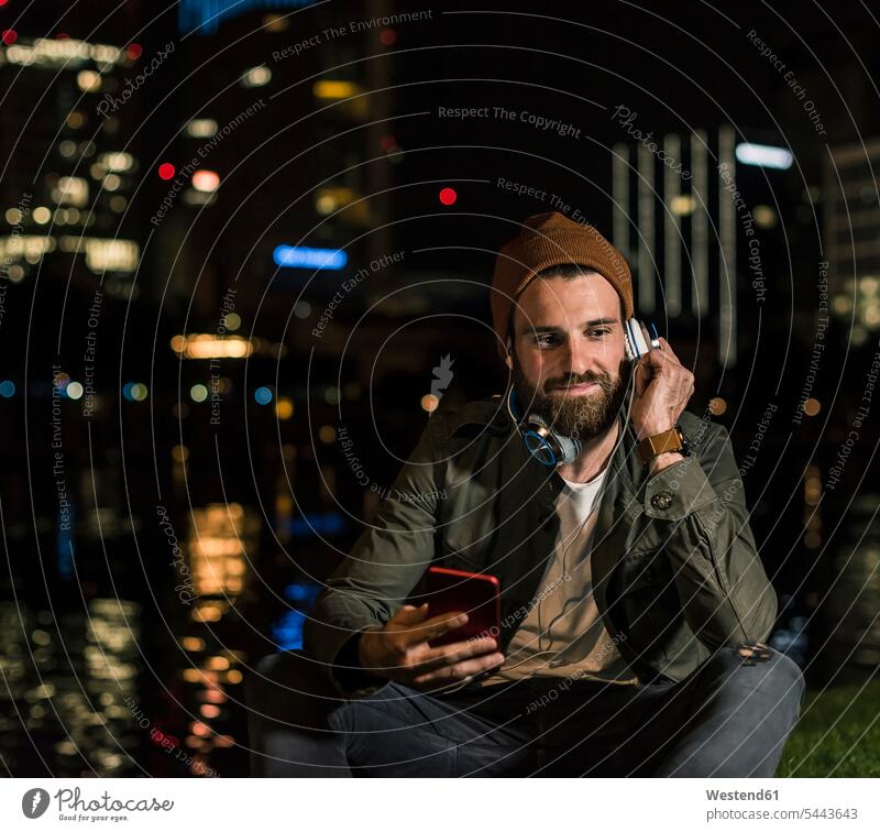Stylish young man with cell phone and headphone sitting at urban riverside at night headphones headset smiling smile by night nite night photography men males