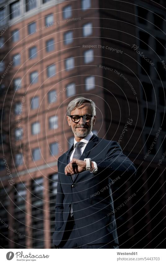 Grey-haired businessman in front of red skyscraper looking at his smartwatch portrait portraits skyscrapers multistory building high rise smart watch smiling