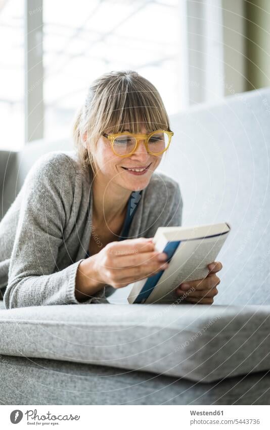 Smiling woman lying on couch reading book books females women laying down lie lying down smiling smile settee sofa sofas couches settees Adults grown-ups