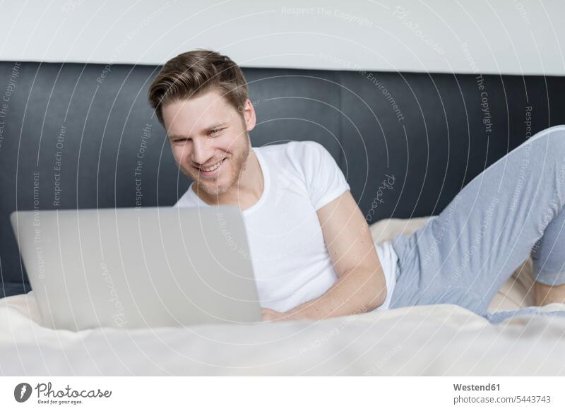 Smiling young man lying on bed using laptop beds smiling smile Laptop Computers laptops notebook men males computer computers Adults grown-ups grownups adult