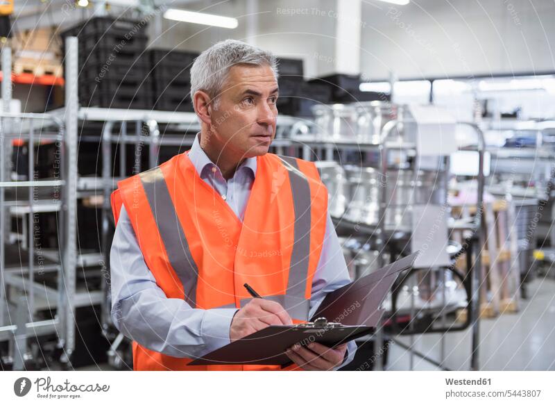 Man in factory hall wearing safety vest holding clipboard man men males scrutiny scrutinizing clipboards clip-board clip-boards clip board working At Work