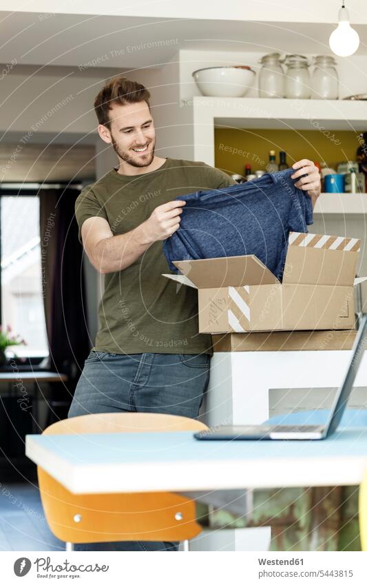 Smiling young man at home unpacking parcel with clothing packet packets parcels smiling smile clothes men males Adults grown-ups grownups adult people persons