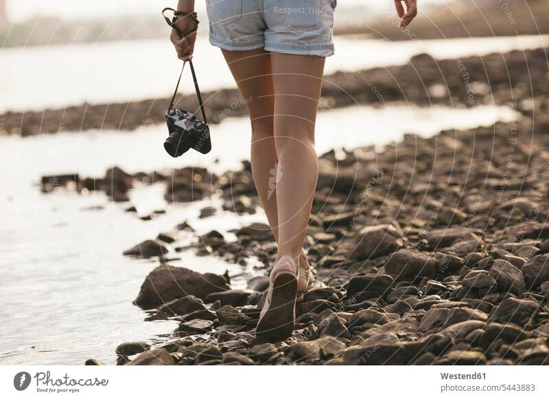 Woman holding a camera walking on stony beach River Rivers going woman females women cameras water waters body of water Adults grown-ups grownups adult people
