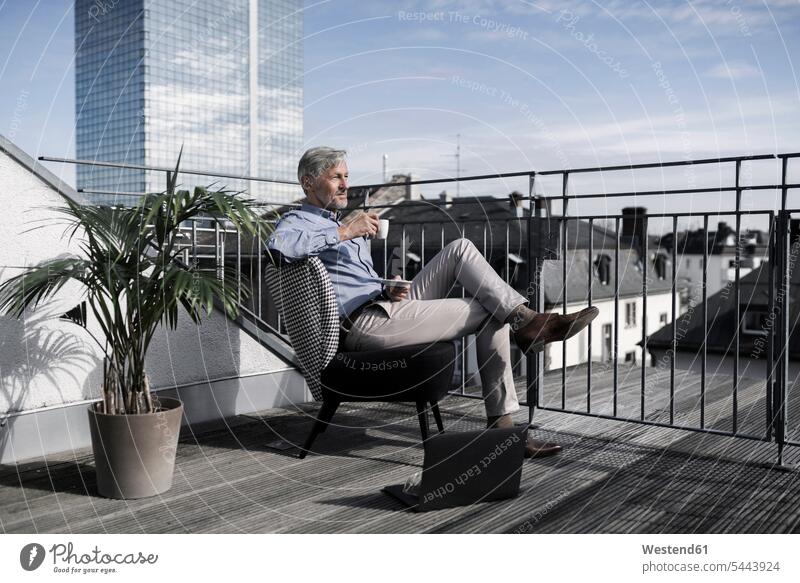 Grey-haired businessman sitting on balcony looking over city and drinking coffee Businessman Business man Businessmen Business men laptop Laptop Computers