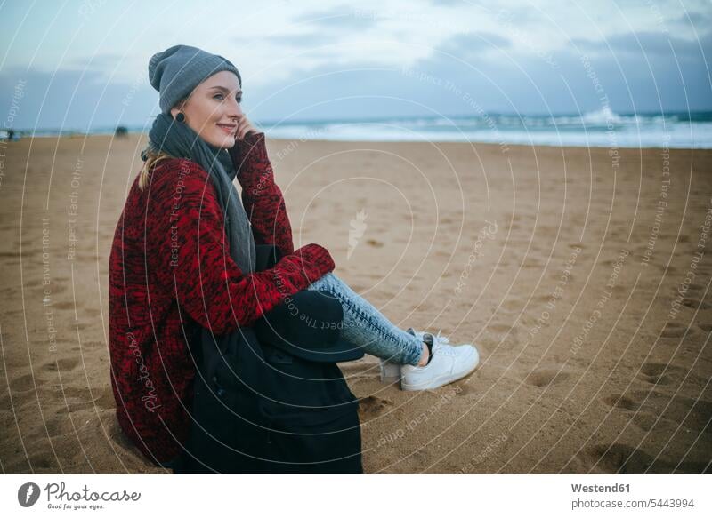 Smiling young woman sitting on the beach in winter beaches Seated females women smiling smile Adults grown-ups grownups adult people persons human being humans
