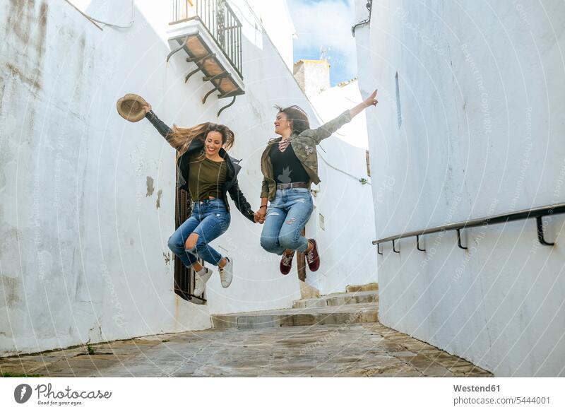 Two happy young women jumping in an alley of a town Fun having fun funny female friends Leaping mate friendship jumps city cities towns woman females lane