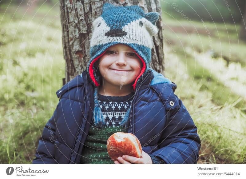 Portrait of grinning boy eating a Berliner in forest boys males woolly hat Wooly Hat Knit-Hat Knit Hats wool cap child children kid kids people persons