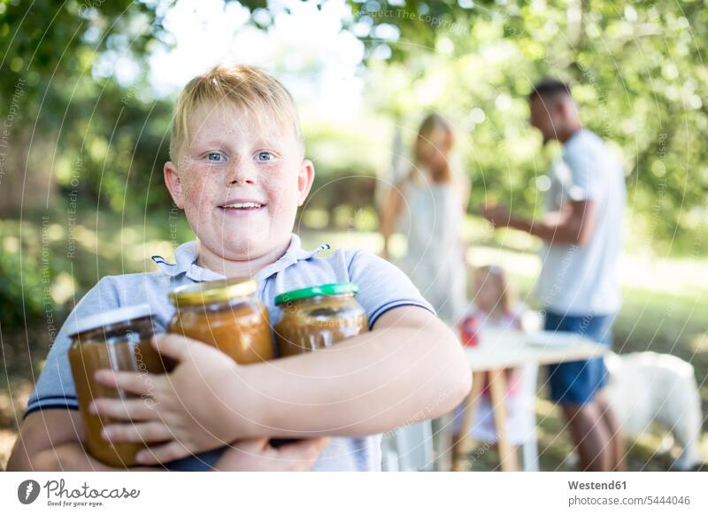 Portrait of boy holding jars outdoors with family in background preserving jar Mason Jars mason jar preserving jars kilner jar preserves jar boys males child