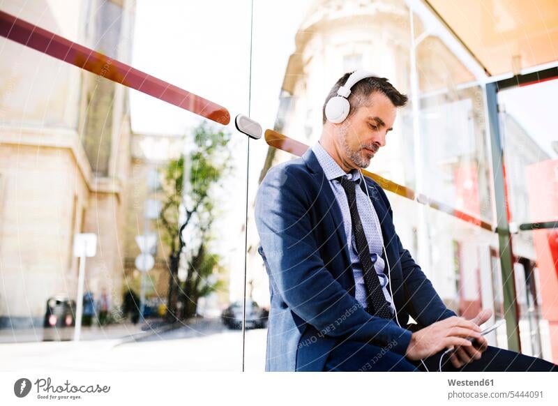 Businessman with smartphone and headphones waiting at the bus stop mobile phone mobiles mobile phones Cellphone cell phone cell phones commuter commuters