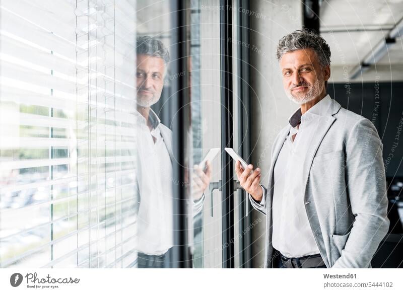 Portrait of smiling mature businessman at the window in office holding cell phone offices office room office rooms smile mobile phone mobiles mobile phones