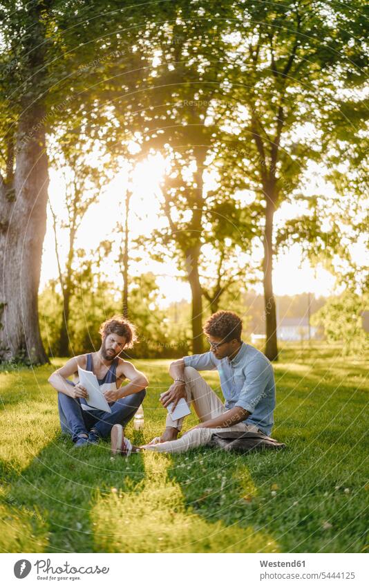 Two friends sitting in a park with mobile device and papers man men males parks mobile phone mobiles mobile phones Cellphone cell phone cell phones documents