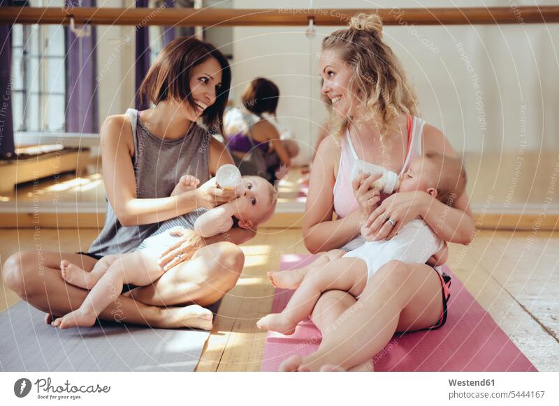 Two mothers bottle-feeding their babies in exercise room mommy ma mummy mama Fun having fun funny exercising training practising smiling smile baby infants