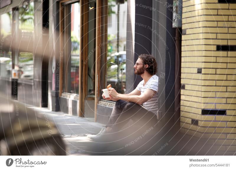 Young man with coffee mug and croissant sitting at entrance of a coffee shop men males Adults grown-ups grownups adult people persons human being humans