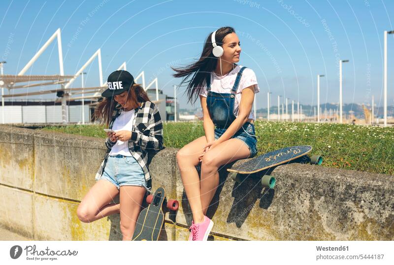 Young woman with headphones listening music while her friend looking at smartphone Longboard female friends female skateboarder female skater
