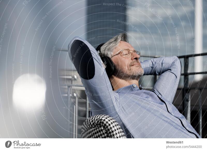 Grey-haired man relaxing in chair on balcony listening to music Businessman Business man Businessmen Business men sitting Seated hearing relaxed relaxation