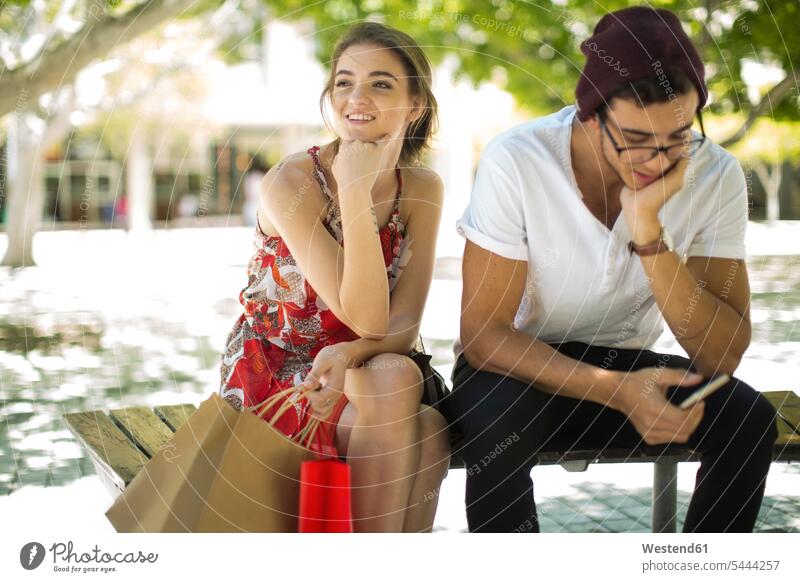 Young couple sitting on bench with shopping bags and cell phone shopping-bag shopping-bags mobile phone mobiles mobile phones Cellphone cell phones twosomes