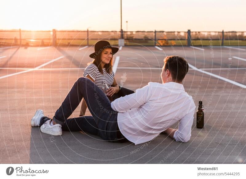 Young couple sitting on parking level at sunset with beer bottles Beer Bottle Beer Bottles Seated twosomes partnership couples sunsets sundown Beers Ale people