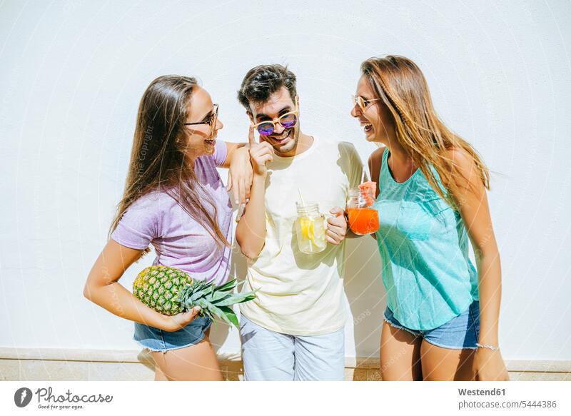 Happy friends holding refreshing drinks and pineapple in front of white wall laughing Laughter happiness happy Drink beverages Drinks Beverage positive Emotion