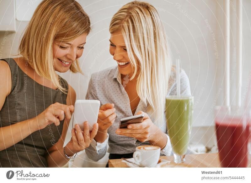 Two happy young women with smartphones in a cafe talking speaking female friends mobile phone mobiles mobile phones Cellphone cell phone cell phones smiling