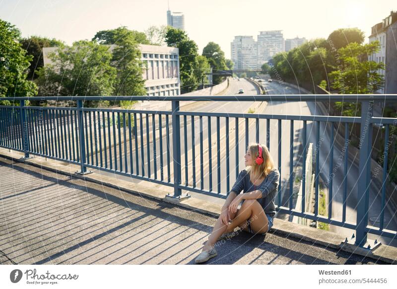 Young woman sitting on bridge listening music with headphones Seated females women hearing headset bridges smiling smile Adults grown-ups grownups adult people