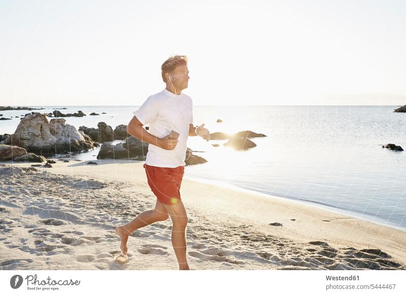 Mature man jogging on the beach while listening music with smartphone and earphones beaches jogger joggers Jogging fitness sport sports Sea ocean men males
