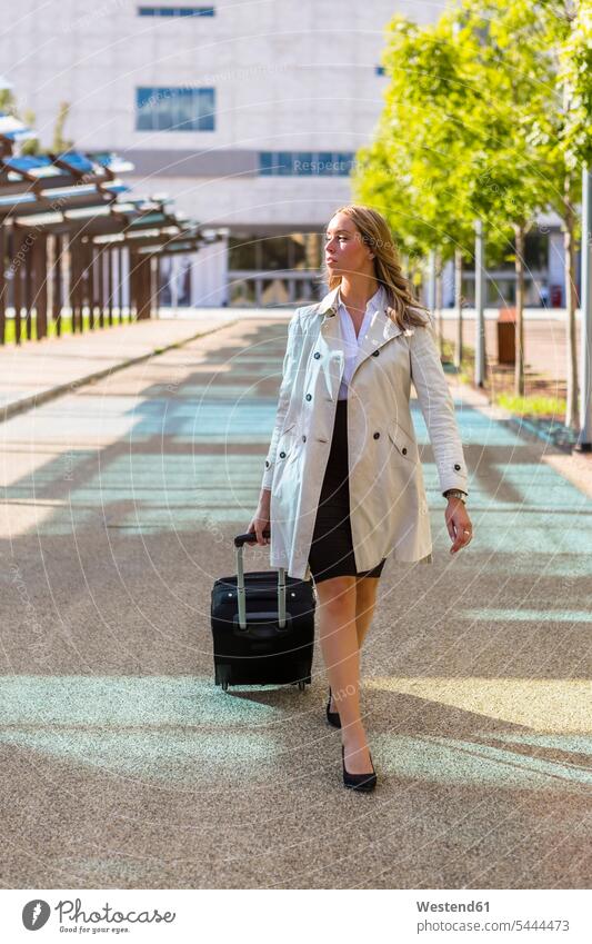 Businesswoman with suitcase wearing trench coat businesswoman businesswomen business woman business women rolling suitcase rolling suitcases business people