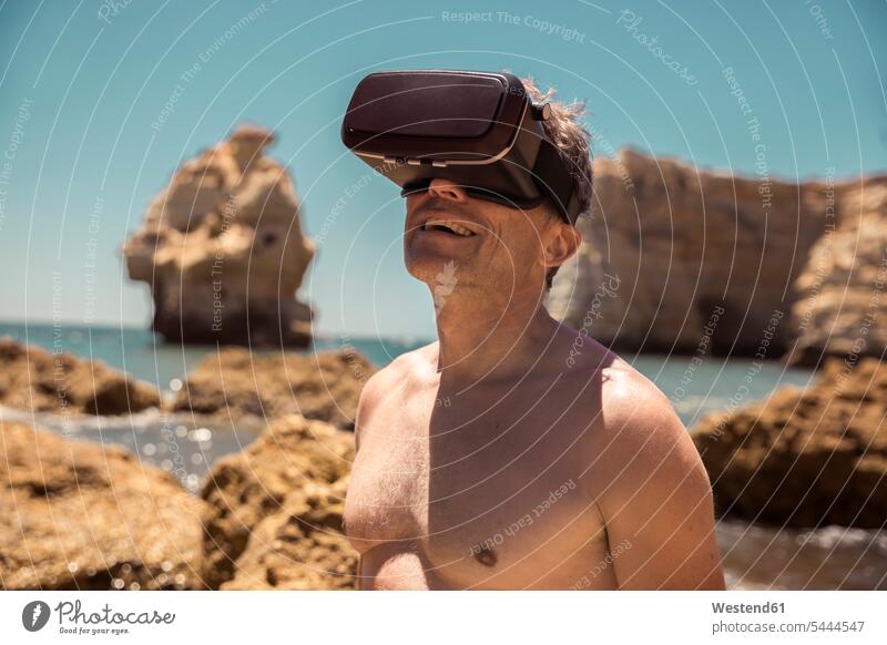 Mature man wearing VR glasses between rocks on beach virtual reality men males beaches specs Eye Glasses spectacles Eyeglasses Adults grown-ups grownups adult