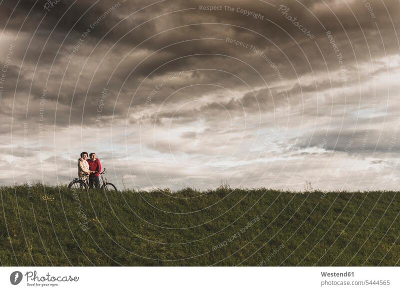 Senior couple with bicycles in rural landscape under cloudy sky embracing embrace Embracement hug hugging senior men senior man elder man elder men