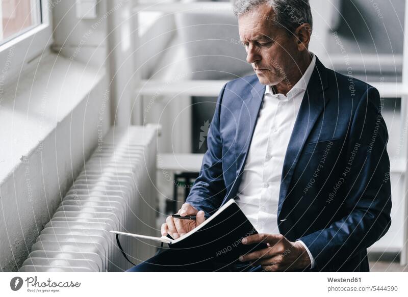 Pensive businessman with notebook sitting beside window notebooks Businessman Business man Businessmen Business men business people businesspeople