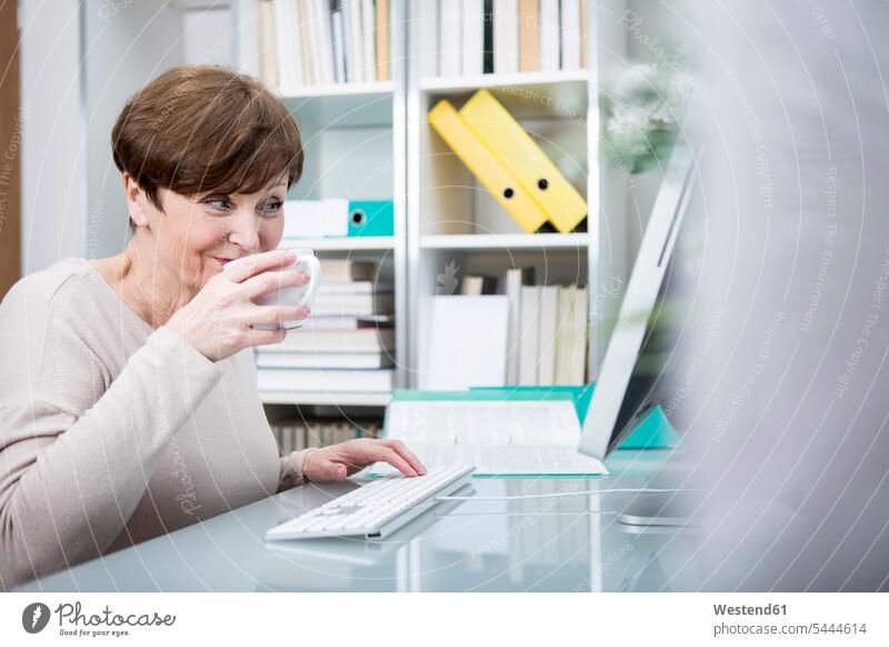 Senior woman working at computer with a cup of coffee females women At Work drinking Coffee sitting Seated learning senior women elder women elder woman old
