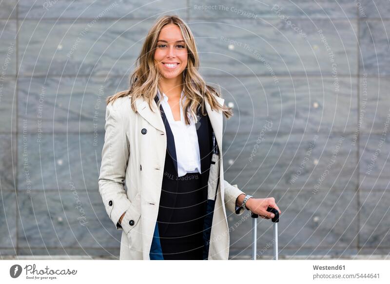 Portrait of smiling businesswoman with suitcase wearing trench coat businesswomen business woman business women portrait portraits rolling suitcase