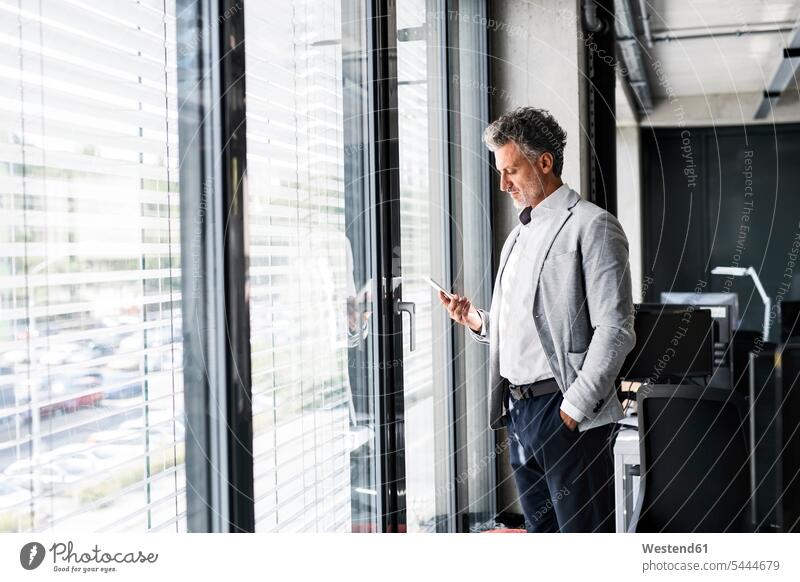 Mature businessman standing at the window in office holding cell phone mobile phone mobiles mobile phones Cellphone cell phones offices office room office rooms
