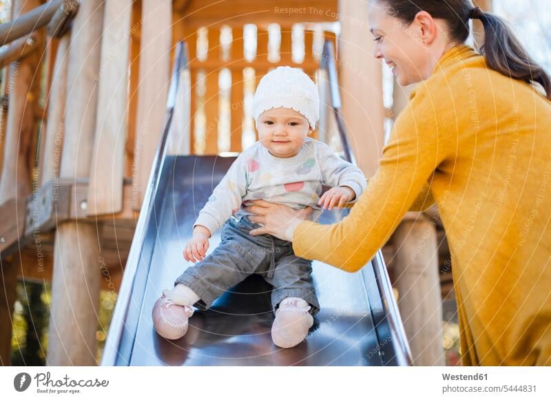 Portrait of baby girl sitting on shute on playground being held by her mother portrait portraits infants nurselings babies slide Chute playground slide slides