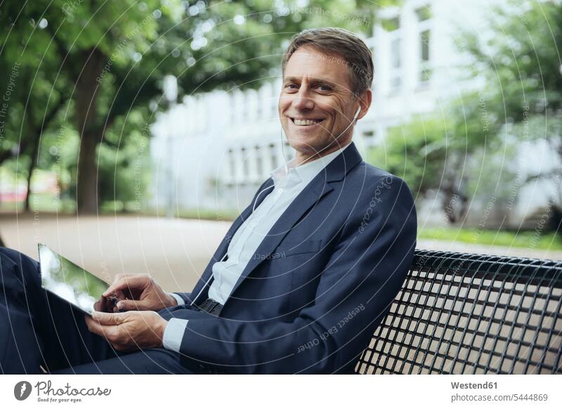 Smiling mature businessman sitting on park bench with tablet and earphones ear phone ear phones smiling smile Businessman Business man Businessmen Business men