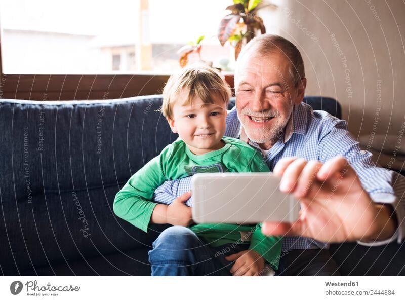 Grandfather and grandson sitting on couch,taking smartphone selfies sharing share Selfie Selfies playing photographing grandfather grandpas granddads