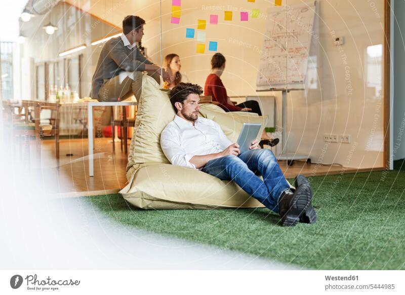 Man in office using tablet in bean bag with meeting in background Post it Post-it offices office room office rooms colleagues man men males Business Meeting