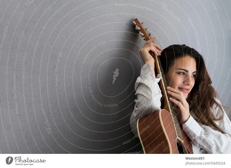 Portrait of smiling young woman with guitar in front of grey background guitars portrait portraits females women stringed instrument stringed instruments