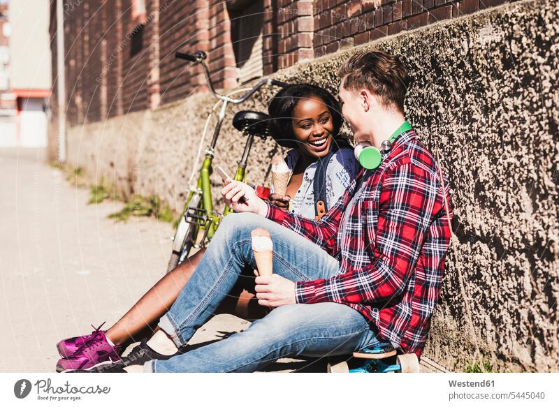 Young couple sitting on ground, eating icecream mobile phone mobiles mobile phones Cellphone cell phone cell phones Seated multicultural twosomes partnership
