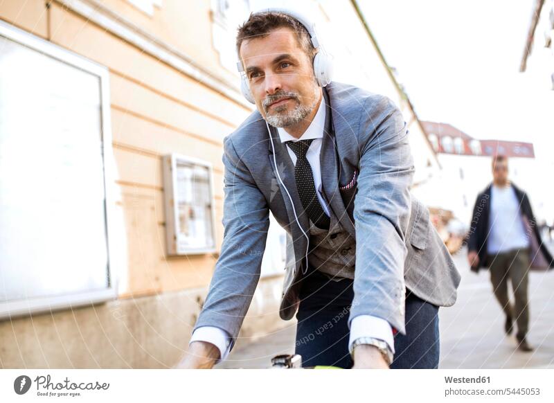 Mature businessman with headphones riding bicycle in the city bikes bicycles men males town cities towns headset Adults grown-ups grownups adult people persons