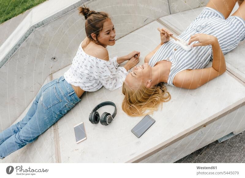 Two happy young women lying on ramp in a skatepark making music female friends laying down lie lying down Skateboard Park skate park Ramp happiness woman