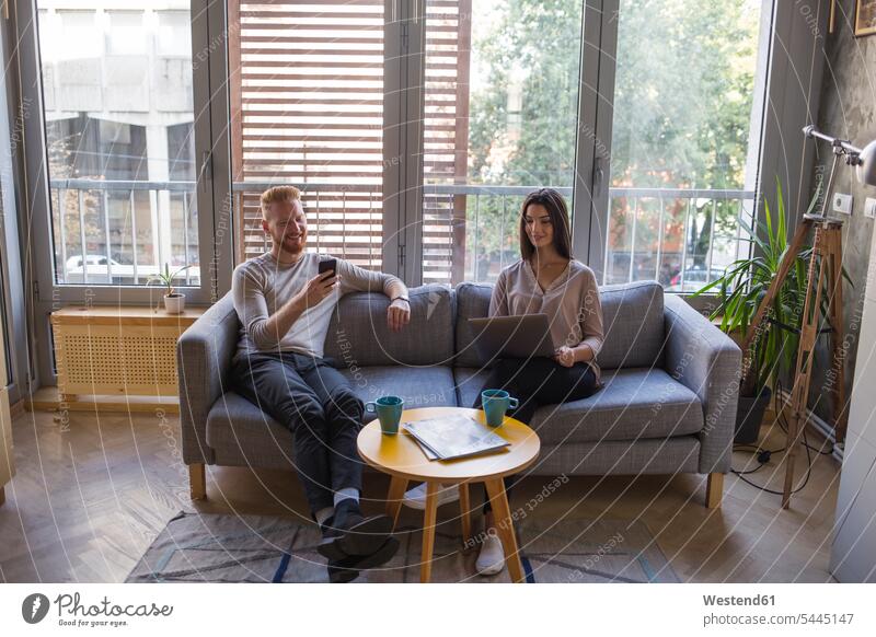 Couple sitting on couch with cell phone and laptop Seated couple twosomes partnership couples settee sofa sofas couches settees looking eyeing mobile phone
