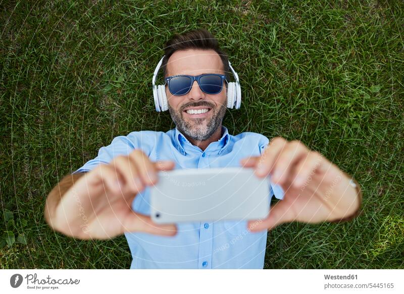 Portrait of laughing man lying on a meadow taking selfie with smartphone, top view Smartphone iPhone Smartphones men males meadows headphones headset Laughter