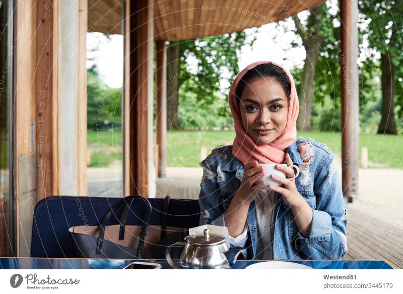 Young woman wearing hijab holding a cup on a terrace of a cafe headscarf head scarf head scarves Head Scarf head cloths headscarves Tea Teas Muslim portrait