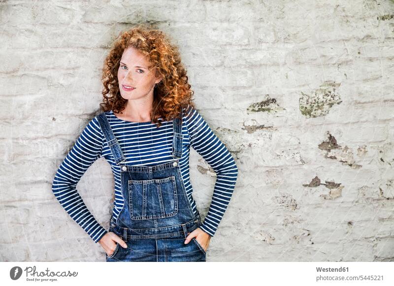 Portrait of smiling young woman wearing denim dungarees leaning against white brick wall females women portrait portraits Adults grown-ups grownups adult people